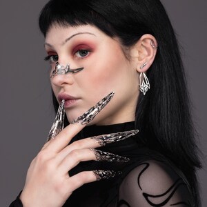 May include: A person wearing a black sheer top with a silver nose ring and silver rings on their fingers. The rings are shaped like claws.