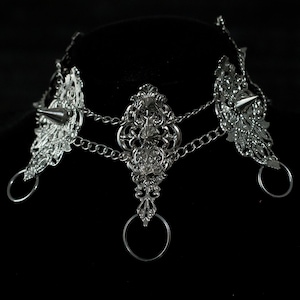 May include: Silver choker necklace with intricate filigree designs and three spikes. The necklace has two chain links and three dangling rings.