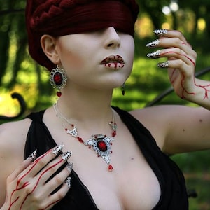 Puede incluir: Una mujer con el pelo rojo y una venda en los ojos lleva un collar de plata con un colgante de piedra roja y pendientes a juego. Sus manos est&aacute;n adornadas con anillos de plata y esmalte de u&ntilde;as rojo.