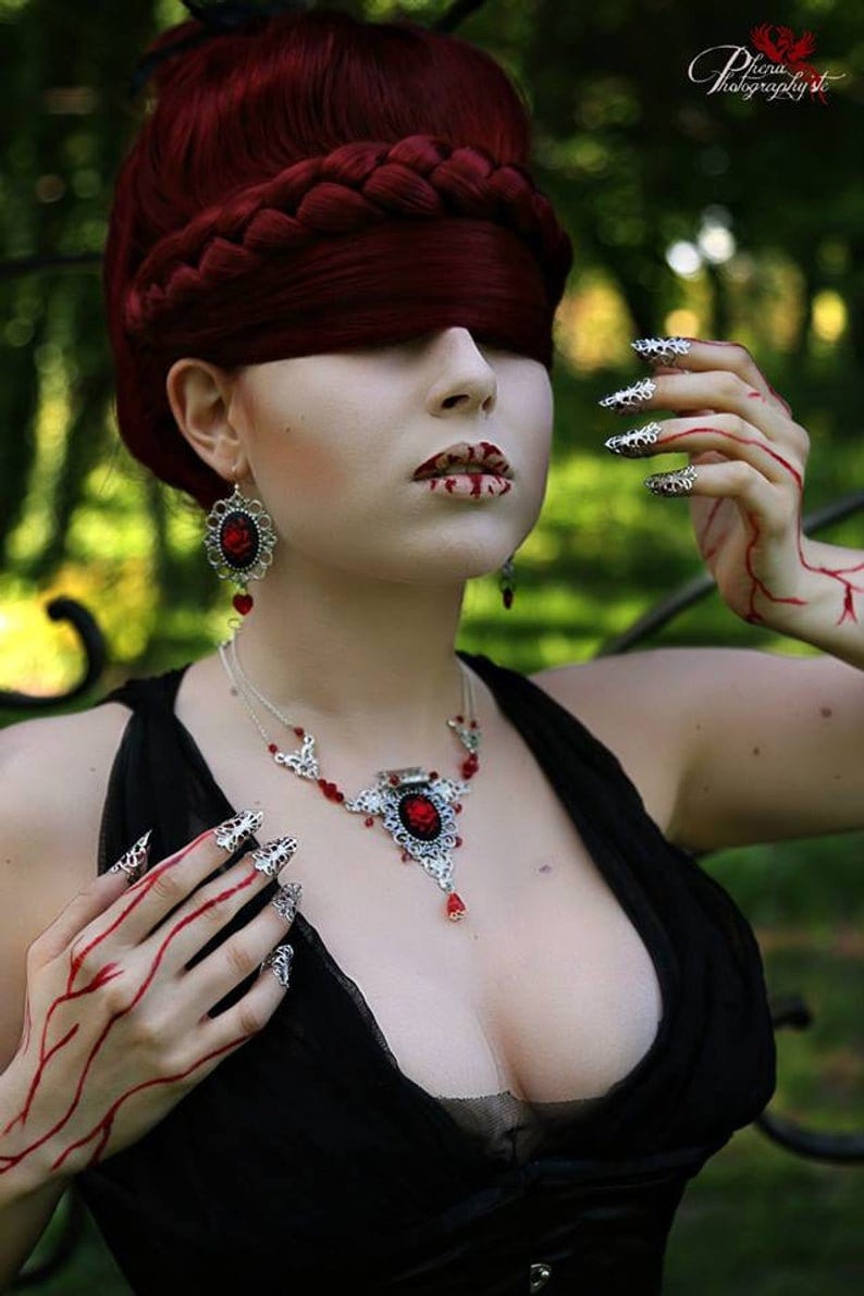 Puede incluir: Una mujer con el pelo rojo y una venda en los ojos lleva un collar de plata con un colgante de piedra roja y pendientes a juego. Sus manos est&aacute;n adornadas con anillos de plata y esmalte de u&ntilde;as rojo.