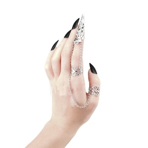 May include: A silver ring with a chain connecting to a second ring. The ring is on a finger with black nail polish.