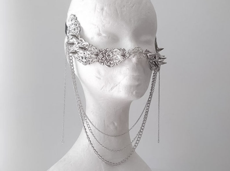 Puede incluir: Una m&aacute;scara facial plateada con un dise&ntilde;o delicado e intrincado y p&uacute;as. La m&aacute;scara est&aacute; unida a una cadena que cuelga del cuello.