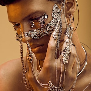 Puede incluir: Una persona con una m&aacute;scara facial plateada y varios anillos plateados en los dedos. La m&aacute;scara es ornamentada y presenta detalles intrincados. La persona tambi&eacute;n lleva un collar y pendientes plateados.