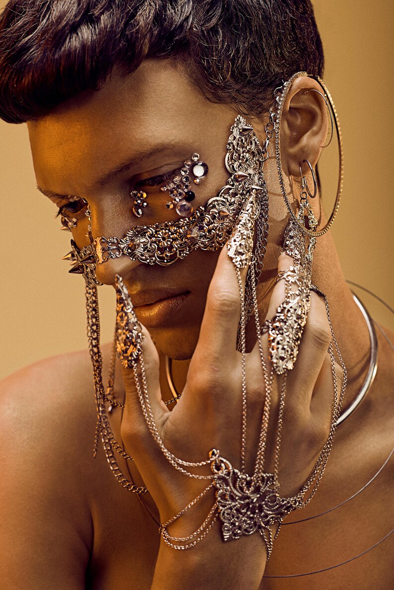 Puede incluir: Una persona con una m&aacute;scara facial plateada y varios anillos plateados en los dedos. La m&aacute;scara es ornamentada y presenta detalles intrincados. La persona tambi&eacute;n lleva un collar y pendientes plateados.