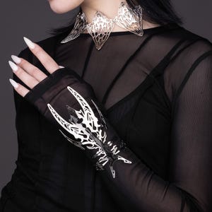 Puede incluir: Un brazalete de mu&ntilde;eca de metal plateado con un dise&ntilde;o detallado e intrincado que se asemeja a una mariposa estilizada. El brazalete se lleva sobre una camisa negra de manga larga y transparente.