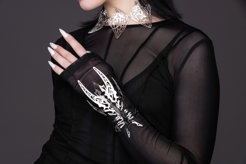 Puede incluir: Un brazalete de mu&ntilde;eca de metal plateado con un dise&ntilde;o detallado e intrincado que se asemeja a una mariposa estilizada. El brazalete se lleva sobre una camisa negra de manga larga y transparente.