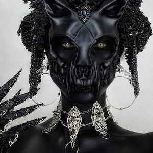 May include: A person wearing a black skull mask with a black and silver beaded headdress and a silver chain necklace. The person is also wearing black gloves with long, sharp nails.