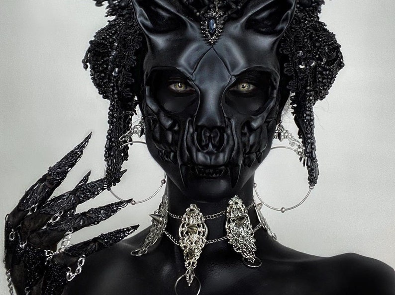 May include: A person wearing a black skull mask with a black and silver beaded headdress and a silver chain necklace. The person is also wearing black gloves with long, sharp nails.