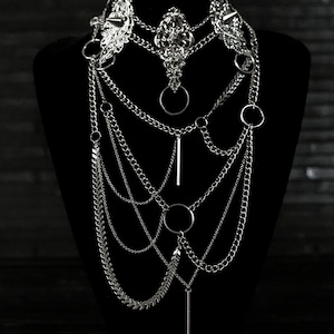 May include: A silver layered necklace with multiple chains, including a chain with arrowhead-shaped links, and several circular and bar-shaped pendants.