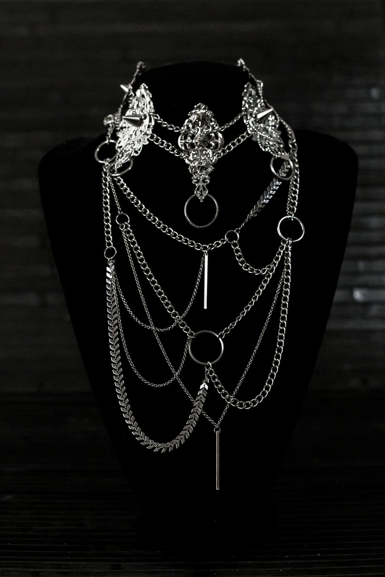 May include: A silver layered necklace with multiple chains, including a chain with arrowhead-shaped links, and several circular and bar-shaped pendants.