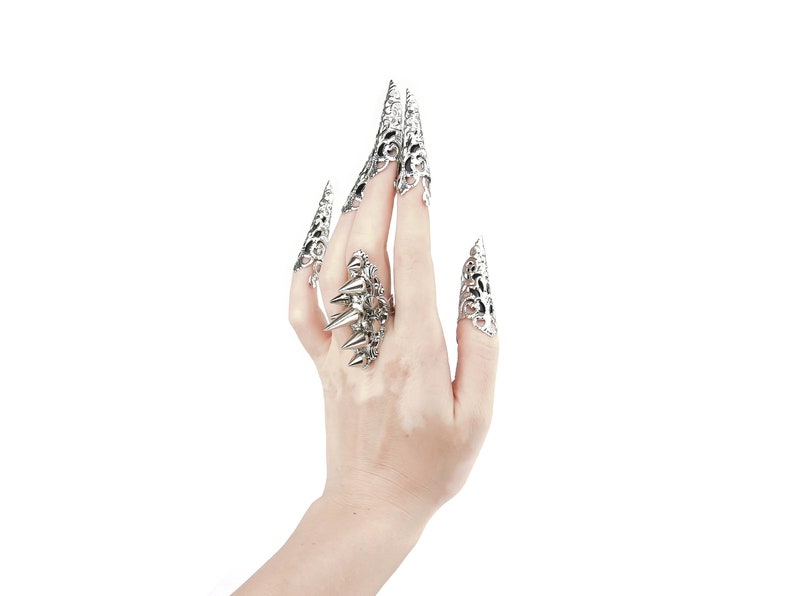 Pu&ograve; includere: Una mano che indossa diversi anelli d'argento con disegni intricati. Gli anelli sono sull'indice, il medio e l'anulare. Gli anelli hanno la forma di artigli.