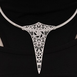 May include: A silver choker necklace with a delicate, intricate pendant featuring a geometric design. The pendant is shaped like a teardrop and has a pointed tip.
