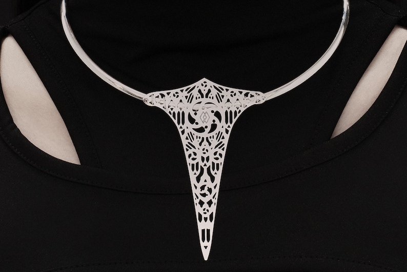 May include: A silver choker necklace with a delicate, intricate pendant featuring a geometric design. The pendant is shaped like a teardrop and has a pointed tip.