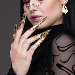 May include: A person wearing gold claw-like rings on their fingers. The rings are intricate and detailed, with a sharp, pointed design.