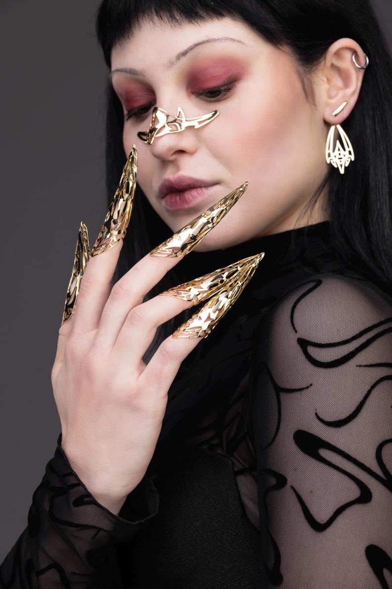 May include: A person wearing gold claw-like rings on their fingers. The rings are intricate and detailed, with a sharp, pointed design.