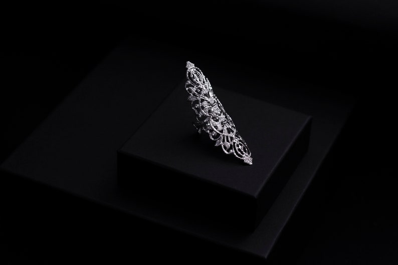 Op de afbeelding: Een zilveren ring met een delicaat, ingewikkeld ontwerp. De ring ligt op een zwarte displaystandaard.