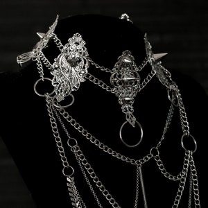 May include: A silver layered chain necklace with ornate metal accents and spikes. The necklace is displayed on a black mannequin bust.