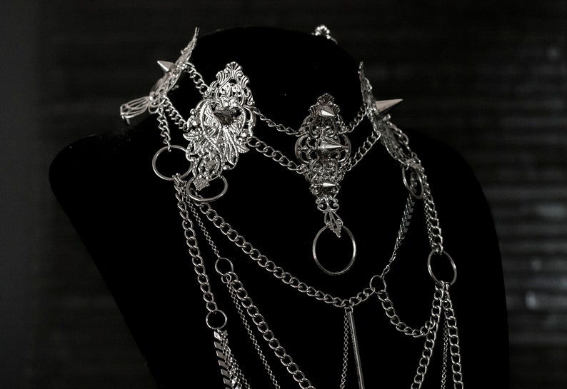 May include: A silver layered chain necklace with ornate metal accents and spikes. The necklace is displayed on a black mannequin bust.