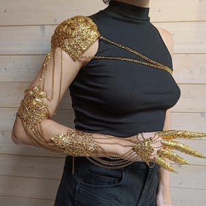 May include: Gold-toned metal arm and hand jewelry with intricate designs. The arm piece features a large, ornate design on the shoulder and extends down the arm with chains and smaller decorative elements. The hand piece features long, claw-like fingers.