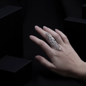 Op de afbeelding: Een zilveren filigraanring met een delicaat, ingewikkeld ontwerp. De ring wordt gedragen op de middelvinger van een hand, met de vingers gestrekt en de palm naar de camera gericht.