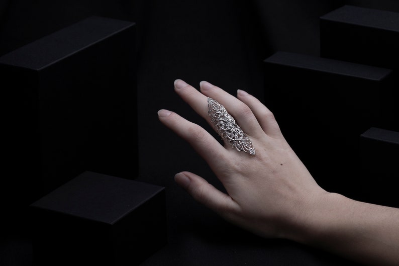 Op de afbeelding: Een zilveren filigraanring met een delicaat, ingewikkeld ontwerp. De ring wordt gedragen op de middelvinger van een hand, met de vingers gestrekt en de palm naar de camera gericht.