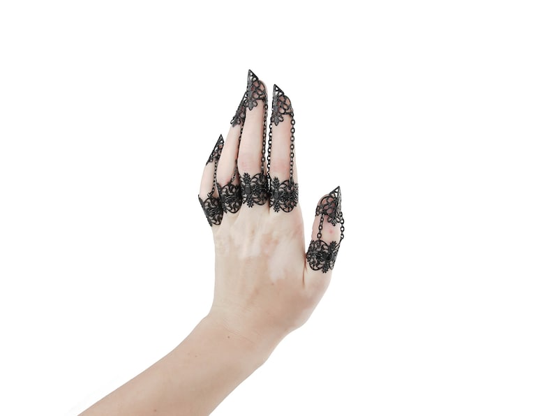 Puede incluir: Una mano con cinco anillos de filigrana negra en cada dedo. Los anillos est&aacute;n conectados por cadenas.