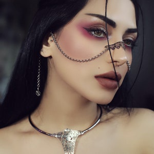 May include: A woman with dark hair and dramatic makeup wears a silver chain nose ring and a silver choker necklace with a large, ornate pendant.