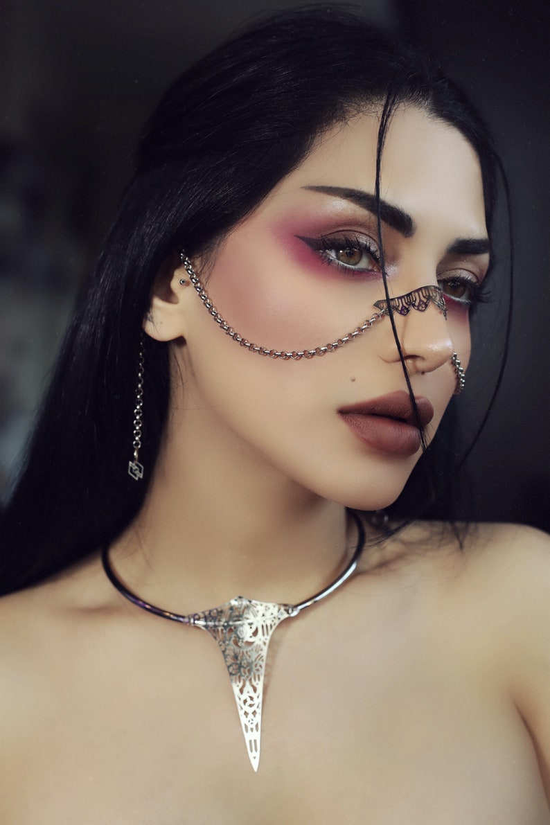 May include: A woman with dark hair and dramatic makeup wears a silver chain nose ring and a silver choker necklace with a large, ornate pendant.