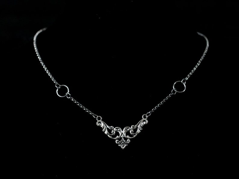 May include: A silver necklace with a delicate, ornate pendant and two small circular links. The pendant features a swirling, floral design.