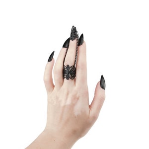 Puede incluir: Un anillo negro con un dise&ntilde;o delicado e intrincado y una peque&ntilde;a piedra negra en el centro. El anillo se lleva en el dedo medio de una mano con u&ntilde;as largas, negras y puntiagudas.