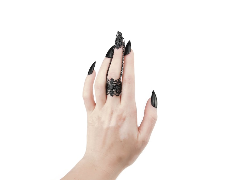 Puede incluir: Un anillo negro con un dise&ntilde;o delicado e intrincado y una peque&ntilde;a piedra negra en el centro. El anillo se lleva en el dedo medio de una mano con u&ntilde;as largas, negras y puntiagudas.