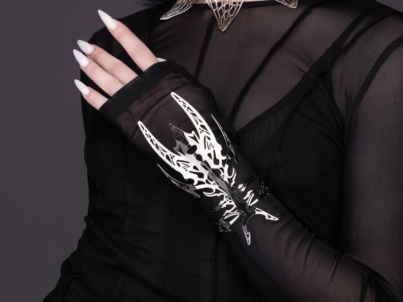Puede incluir: Guantes sin dedos de malla negra transparente con un dise&ntilde;o decorativo de metal plateado en el dorso de la mano. El dise&ntilde;o es intrincado y presenta un patr&oacute;n geom&eacute;trico.