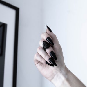 May include: A black ring with a detailed design on the top. The ring is worn on the middle finger of a hand with long, black, pointed nails.