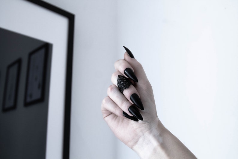 May include: A black ring with a detailed design on the top. The ring is worn on the middle finger of a hand with long, black, pointed nails.