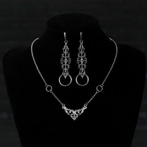 May include: A silver necklace and earrings set with a delicate, ornate design. The necklace features a V-shaped pendant with a small heart at the bottom. The earrings are long and dangle, with a circular ring at the bottom.