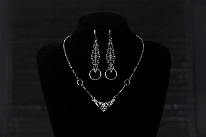 May include: A silver necklace and earrings set with a delicate, ornate design. The necklace features a V-shaped pendant with a small heart at the bottom. The earrings are long and dangle, with a circular ring at the bottom.