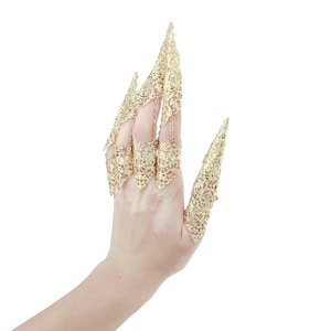 May include: A hand wearing a set of gold, ornate finger rings. The rings are designed to resemble claws or talons, with intricate filigree details.