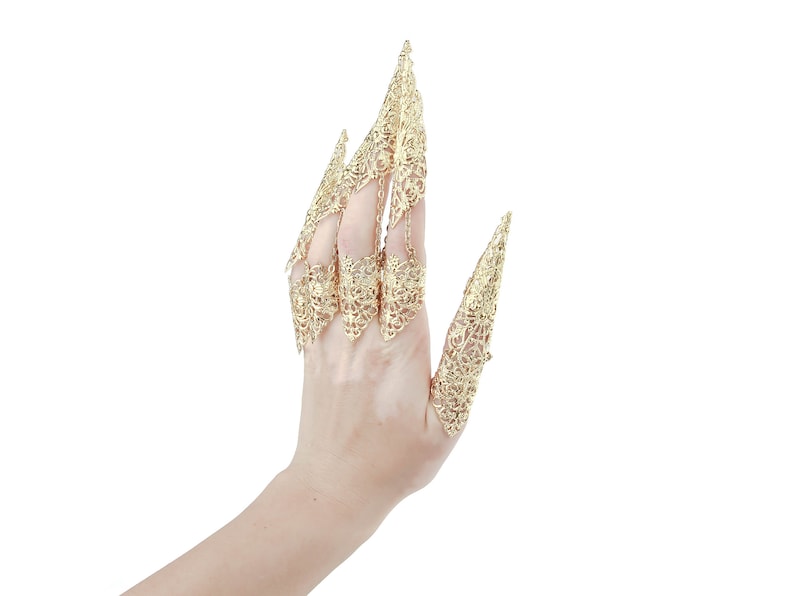 May include: A hand wearing a set of gold, ornate finger rings. The rings are designed to resemble claws or talons, with intricate filigree details.