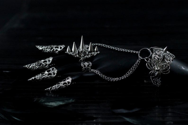 Pu&ograve; includere: Un set di anelli per dita argentati con disegni intricati, tra cui un anello con pi&ugrave; punte. Gli anelli sono collegati da una catena argentata.