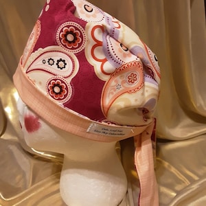 May include: A pink and red paisley patterned surgical scrub hat with a pink tie. The hat has a label that reads "Deli's Sweet Gifts Etsy Shop Delicious".