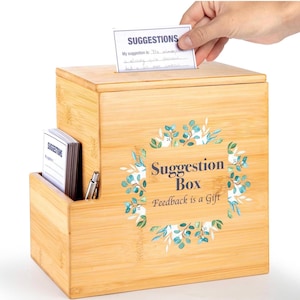 May include: A light brown bamboo suggestion box with a slot on top and a side compartment holding a stack of cards and a silver pen. The front features a floral wreath graphic surrounding the text "Suggestion Box" and "Feedback is a Gift".