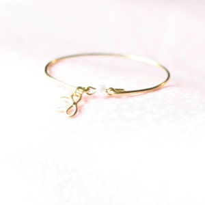 Puede incluir: Una delicada pulsera de brazalete dorada con un pequeño dije de lazo y una sola perla blanca. La pulsera delgada y elegante es perfecta para el uso diario. El dije de lazo añade un toque de fantasía al diseño.