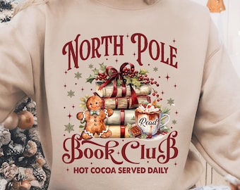 May include: A beige sweatshirt with a Christmas-themed design featuring a gingerbread man, a stack of books, a mug of hot cocoa, and the text "North Pole Book Club + Hot Cocoa Served Daily".
