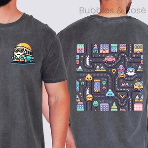 May include: Two charcoal gray t-shirts with different graphic designs. The left shirt features a cartoon van with a smiling face and sunglasses. The right shirt features a colorful cartoon city scene with buildings, roads, and characters.