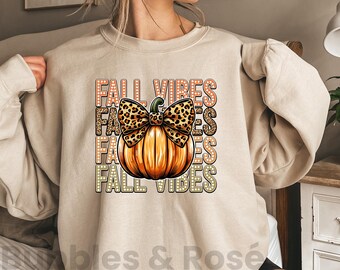 May include: Sand-colored sweatshirt featuring a large orange pumpkin with a leopard print bow. The words "FALL VIBES" are stacked vertically in a retro font. The sweatshirt is a cozy fall-themed apparel item.