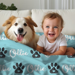 May include: A light blue blanket with a paw print pattern and the name "Ollie" written in white. A dog and a baby are sitting on the blanket.