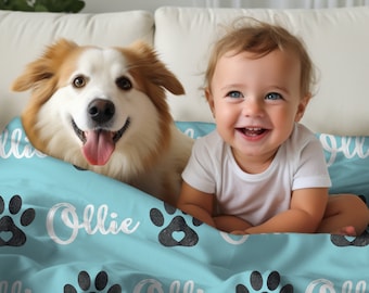 May include: A light blue blanket with a paw print pattern and the name "Ollie" written in white. A dog and a baby are sitting on the blanket.