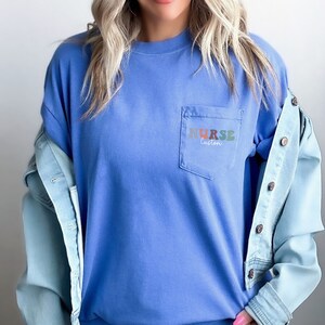 May include: A blue long-sleeved t-shirt with a pocket on the left side. The pocket has the word "NURSE" written in colorful letters with the word "custom" underneath.