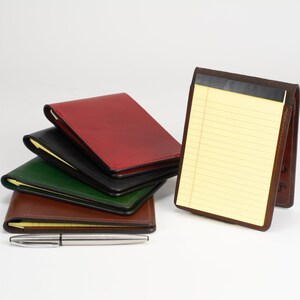 May include: A stack of four leather notepad holders in red, black, green, and brown, with one brown leather holder standing upright containing a yellow lined notepad, accompanied by a silver metal pen placed horizontally in the foreground.