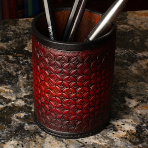 May include: A cylindrical desk organizer made of red leather with a geometric pattern. The top and bottom edges are trimmed with black leather. Pencils and pens are visible inside the organizer.